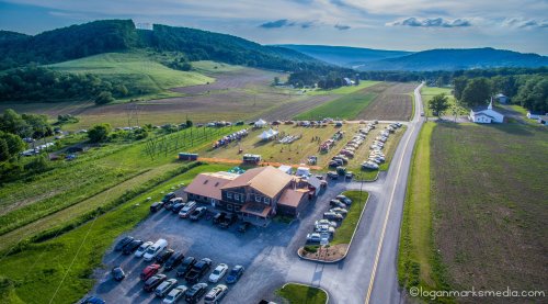 Aerial view of a rural area with a large building, parked cars, fields, and hills in the background.