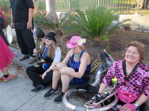 Three women sit on the ground, one holding a tuba, wearing colorful outfits and accessories, surrounded by greenery.