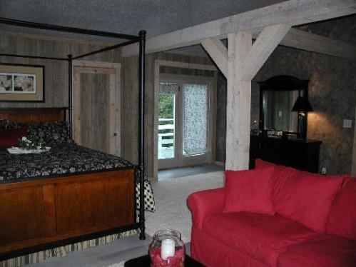 Cozy bedroom with a four-poster bed, red couch, and wooden beams, featuring a view of a balcony through sheer curtains.