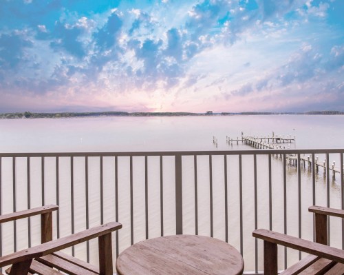 A serene waterfront view from a balcony, featuring wooden chairs and a distant pier under a colorful sky.