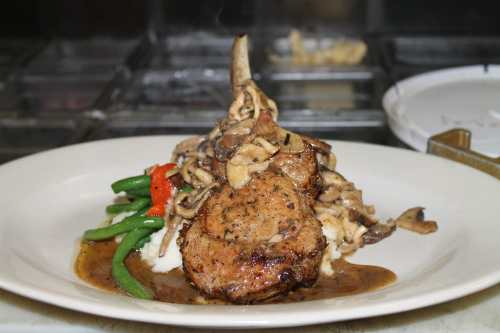 A plated dish featuring a pork chop on mashed potatoes, topped with mushrooms, green beans, and a drizzle of sauce.