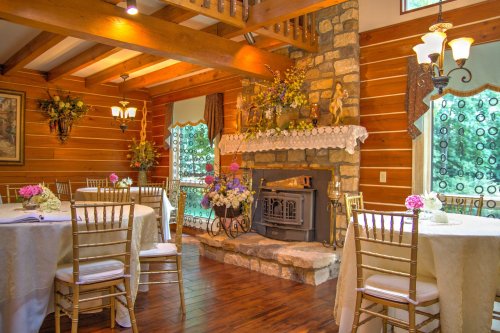 Cozy dining area with wooden beams, stone fireplace, and elegantly set tables adorned with flowers.
