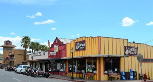 Colorful storefronts line a street, featuring cafes and shops, with motorcycles parked outside under a clear blue sky.
