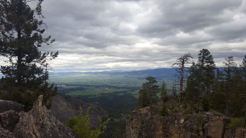 A panoramic view of a valley surrounded by mountains, with cloudy skies and scattered trees in the foreground.
