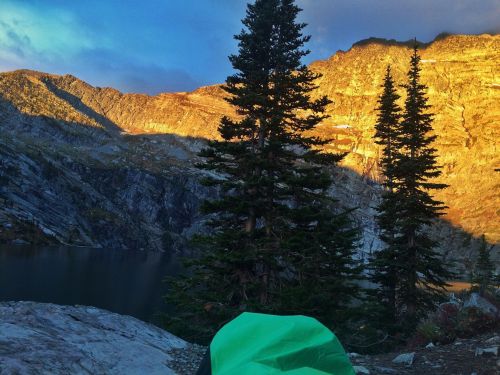 A serene mountain landscape at sunset, featuring tall trees and a calm lake surrounded by rocky terrain.