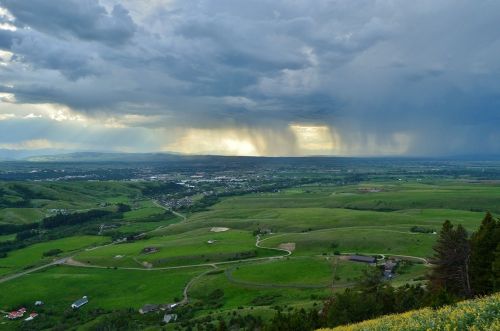 A panoramic view of green hills and a valley under a cloudy sky, with rain falling in the distance.