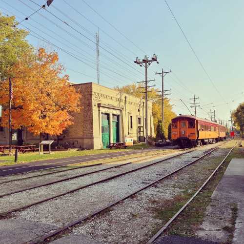 An orange train parked beside a vintage station, with autumn trees and power lines in the background.