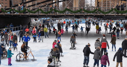 A bustling ice skating rink filled with people of all ages enjoying skating under a bridge decorated for the holidays.