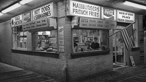 A vintage food stand with signs for hamburgers, hot dogs, and fried chicken, featuring a person behind the counter.