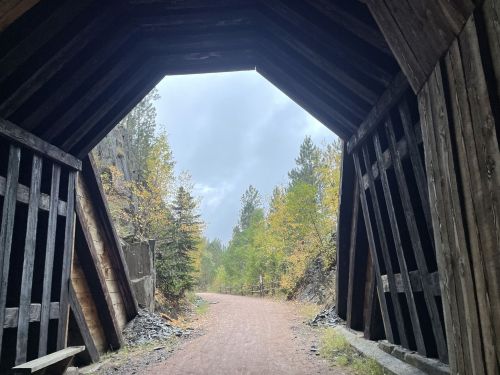 View through a wooden archway leading to a gravel path surrounded by trees with autumn foliage under a cloudy sky.