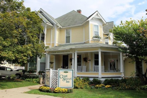 A charming yellow Victorian inn with a porch, surrounded by greenery and flowers, featuring a sign that reads "Grant Street Inn."