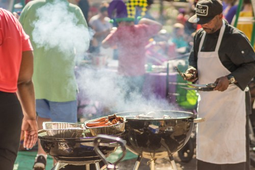 A man grills food at an outdoor event, with smoke rising and people mingling in the background.
