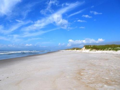 A serene beach scene with soft sand, gentle waves, and a clear blue sky dotted with clouds.