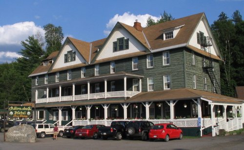 Historic hotel with a green exterior, multiple stories, and a porch, surrounded by trees and parked cars.