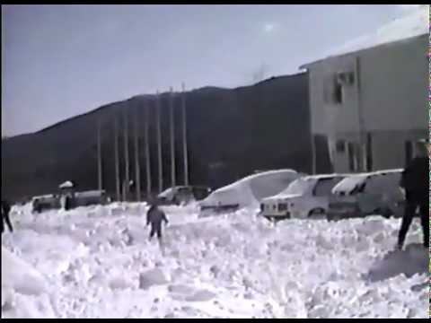 A snowy scene with people playing in deep snow near parked cars and buildings, surrounded by mountains.