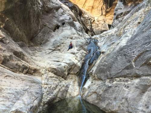 A person sits on a rocky ledge beside a small waterfall in a narrow canyon surrounded by towering stone walls.