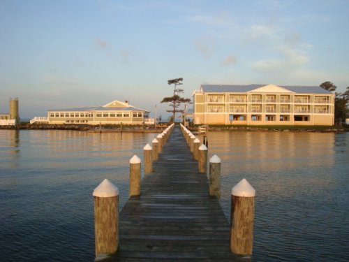 A wooden pier extends into calm water, flanked by two buildings under a soft sunset sky.