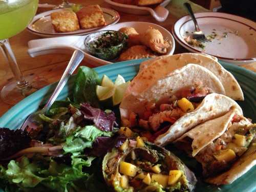 A colorful plate featuring tacos with mango, a side salad, and appetizers in the background, with a drink nearby.