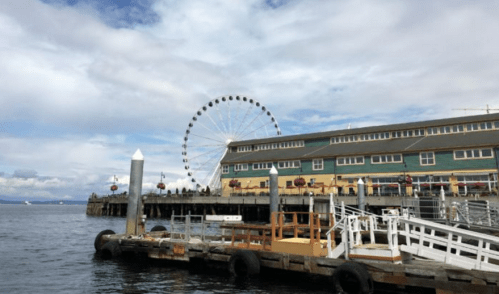 A waterfront view featuring a pier, a large Ferris wheel, and a building with outdoor seating under a cloudy sky.