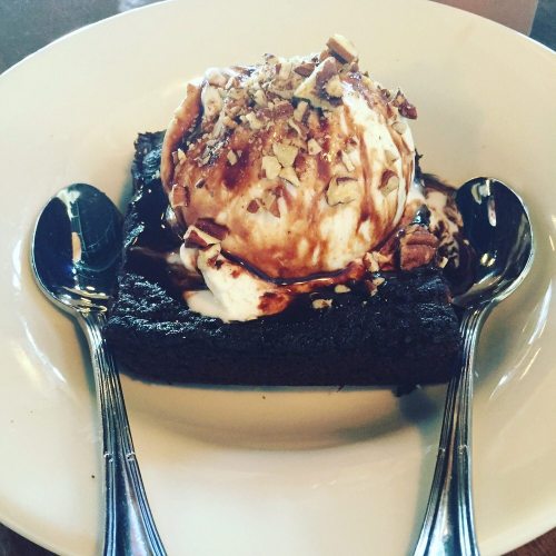 A warm brownie topped with vanilla ice cream, chocolate sauce, and chopped nuts, served with two spoons.