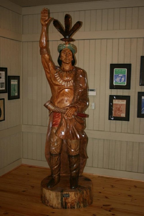 A wooden statue of a Native American figure, raising one hand, adorned with feathers and traditional attire, in a gallery.