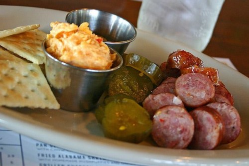 A plate with crackers, pimento cheese, pickles, and sliced sausage in metal cups.