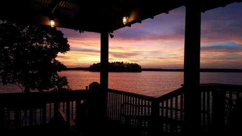 Silhouette of a person on a deck overlooking a tranquil lake at sunset, with vibrant colors in the sky.