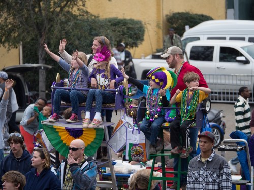 A festive parade scene with children and adults on floats, wearing colorful beads and costumes, waving to the crowd.