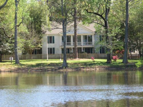 A large white house with green shutters, surrounded by trees, near a calm pond with a wooden fence in the background.