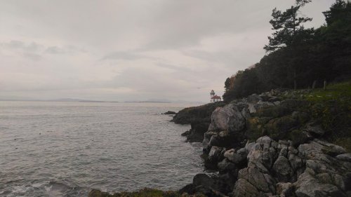 A rocky shoreline with a lighthouse in the distance under a cloudy sky. Calm waters stretch out to the horizon.