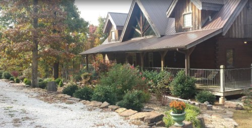 A rustic wooden house with a metal roof, surrounded by trees and a gravel path, featuring a garden in front.