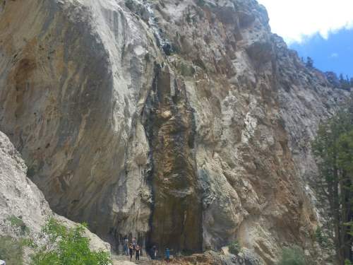 A rocky cliff with a small waterfall cascading down, surrounded by trees and a few people at the base.