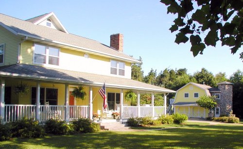 A yellow two-story house with a porch, American flag, and a smaller yellow building in a green yard.