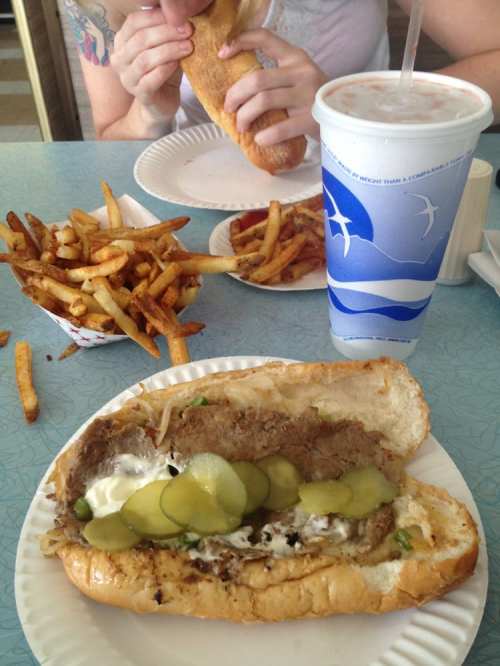 A plate with a sandwich filled with pickles, fries, and a drink in the background.