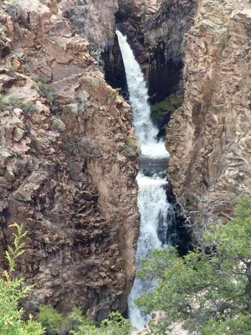 A waterfall cascades between steep, rocky cliffs, surrounded by greenery and rugged terrain.