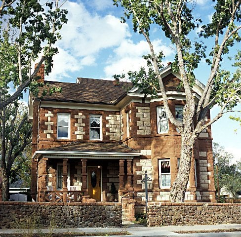 A historic two-story house with stone exterior, large windows, and a porch, surrounded by tall trees and a stone wall.