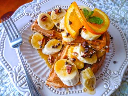 A plate of waffles topped with banana slices, orange slices, pecans, chocolate chips, and syrup, garnished with mint.