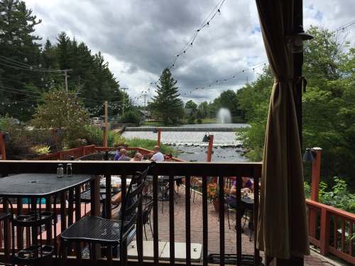View from a deck overlooking a pond with a fountain, surrounded by trees and outdoor seating. Cloudy sky above.