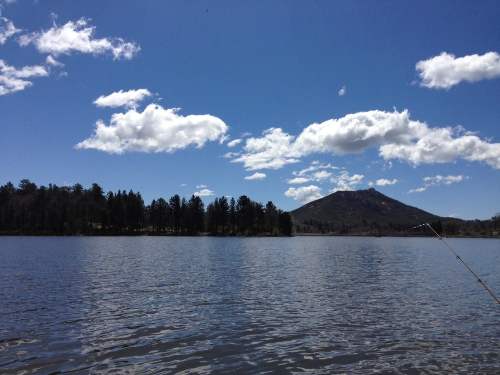 A serene lake scene with a clear blue sky, fluffy clouds, and a mountain in the background, surrounded by trees.