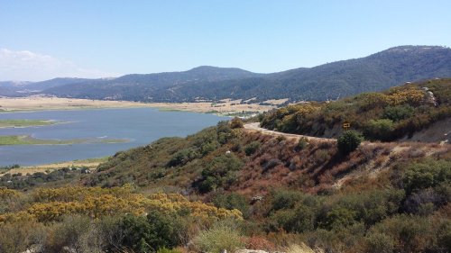 A scenic view of rolling hills and a lake under a clear blue sky, with greenery and a winding road in the foreground.