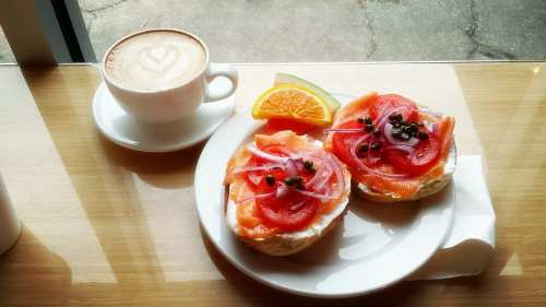 A plate with two bagels topped with smoked salmon, tomatoes, onions, and capers, alongside a cup of coffee and an orange slice.