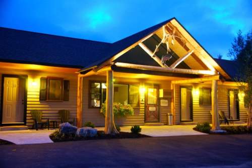A cozy lodge with a wooden entrance, illuminated at dusk, featuring a porch and landscaped surroundings.