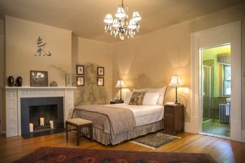 Cozy bedroom with a chandelier, fireplace, and a bed, featuring warm tones and elegant decor. Doorway leads to a bathroom.