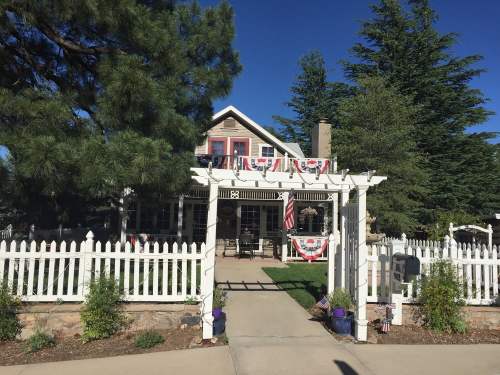A charming house with a white picket fence, decorated with flags and bunting, surrounded by tall trees and a clear blue sky.