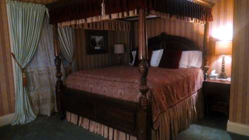 A cozy bedroom featuring a four-poster bed with rich bedding, curtains, and a bedside lamp in a warmly decorated room.