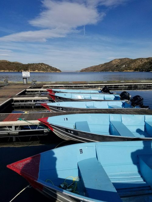 Rowboats lined up on a dock by a calm lake, with hills and a blue sky in the background.