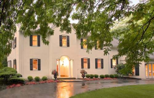 A large, elegant home with white walls, black shutters, and a lush garden, surrounded by trees and a wet driveway.