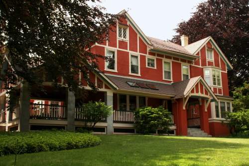 A large red house with white trim, featuring a porch and surrounded by green grass and trees.