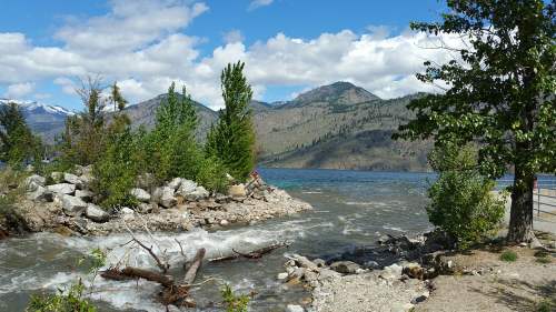 A scenic view of a lake surrounded by mountains, with trees and rocky shores under a partly cloudy sky.