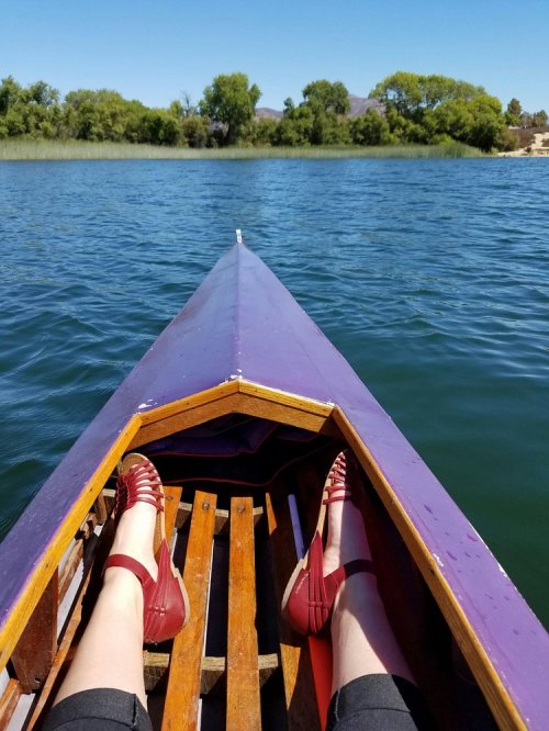 A view from a boat showing feet in red sandals resting on a wooden seat, with calm water and trees in the background.